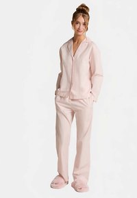 Lichtroze pyjamaset van zachte stof met een overhemd met knopen en broek met elastische taille, aangevuld met zachte roze slippers.