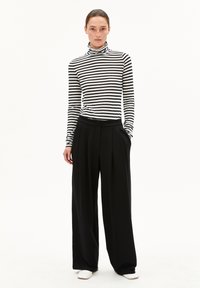 Schwarz-weiß gestreifter Rollkragenpullover kombiniert mit weit geschnittenen schwarzen Hosen, die ein plissiertes Design aufweisen, und weißen Slip-On-Schuhen. Minimalistischer Stil.