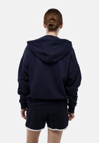 Hoodie bleu marine avec une grande capuche ajustable, des épaules tombantes et une coupe décontractée. Assorti à un short noir avec des bordures blanches.
