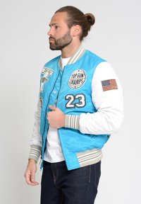 Blauwe bomberjack met witte mouwen, voorzien van geborduurde patches, waaronder "TOP GUN CHAMPS" en de Amerikaanse vlag. Gemêleerde grijze strepen op de manchetten.