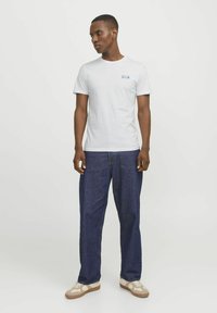 Hvid bomulds T-shirt med et lille blåt logo på venstre bryst, parret med mørkeblå denim jeans. Modellen står foran en neutral baggrund.