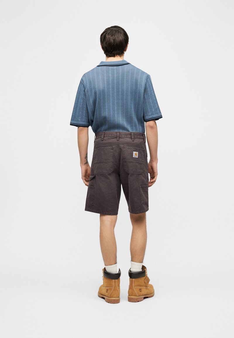 Man draagt een blauw gebreid shirt met korte mouwen, donkerbruine cargoshorts, witte sokken en zandkleurige werkschoenen, staand met de rug naar voren op een witte achtergrond.