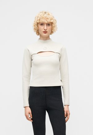 Frau mit lockigem blondem Haar, die ein beigefarbenes langärmeliges Oberteil mit Ausschnittdetail und dunkle, hoch taillierte Hosen trägt, steht vor einem einfarbigen Hintergrund.