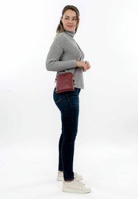 Pequeña bolsa de cuero burdeos cruzada con un diseño elegante y correa ajustable, combinada con un suéter gris de canalé y jeans oscuros.
