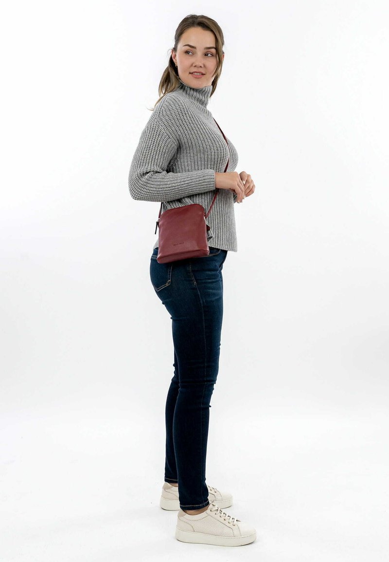 Pequeña bolsa de cuero burdeos cruzada con un diseño elegante y correa ajustable, combinada con un suéter gris de canalé y jeans oscuros.