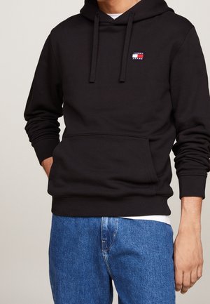 Muž v černé mikině Tommy Jeans s kapucí a přední kapsou na klokaní kapsu, modré džíny, ruce částečně v kapsách, béžové pozadí.