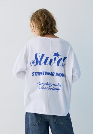 Vrouw met kort golvend haar, gekleed in een witte oversized trui met blauwe tekst en sterrenafbeelding, gecombineerd met losse blauwe spijkerbroek, van achteren gezien.