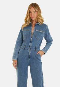 Salopette in denim azzurro chiaro con zip frontale, due tasche sul petto, maniche lunghe e vita con cintura, caratterizzata da un fit rilassato e un tessuto resistente.