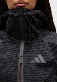 Άτομο που φοράει μαύρο μπουφάν με ψηλό γιακά και φερμουάρ στο μπροστινό μέρος, με διακριτικό ριγέ μοτίβο, εμφανίζοντας μέρος του προσώπου και των χειλιών.