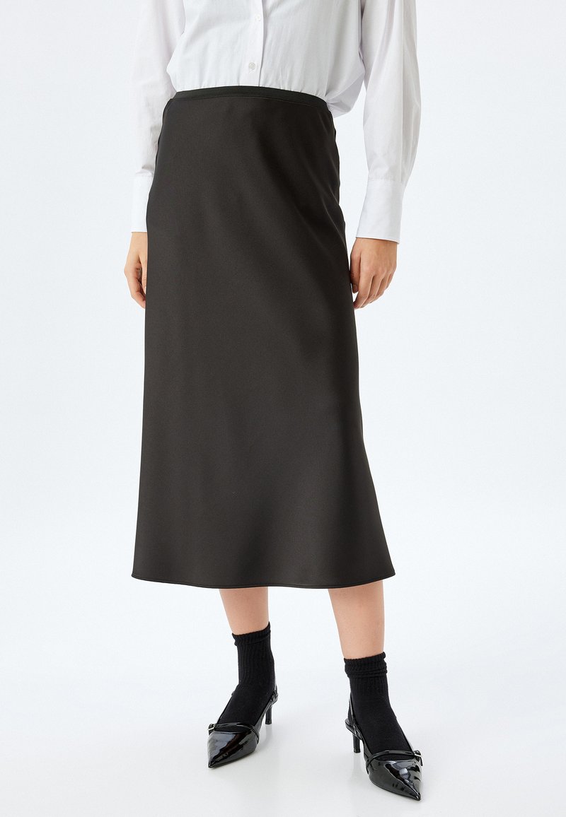 Falda midi negra hecha de tela suave, con una cinturilla ajustada y un ligero corte en A. Combinada con zapatos de tacón negros y calcetas.
