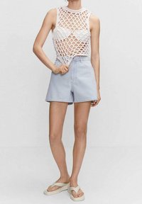Bílý háčkovaný top bez rukávů s diamantovým vzorem, který je v kombinaci se světle modrými a bílými pruhovanými šortkami s vysokým pasem. Na nohou béžové žabky.