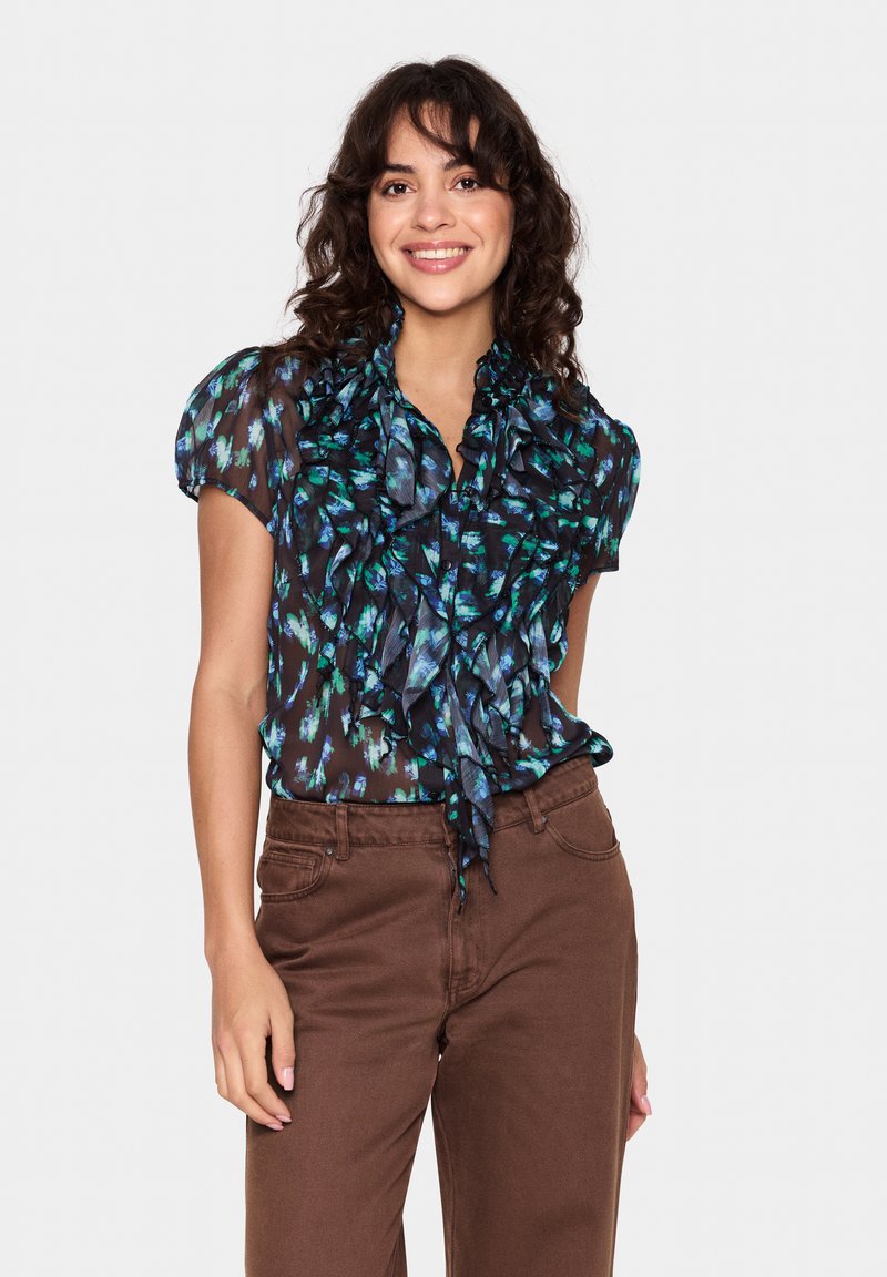 Črna bluzica z zelenim in modrim abstraktnim cvetličnim vzorcem, nabrano ovratnikom, kratkimi napihnjenimi rokavi in zavezanim detajlom okoli vratu. V kombinaciji z rjavimi hlačami.