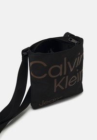 Svart tyg axelväska med dragkedjestängning, med stora bruna bokstäver "Calvin Klein Jeans" på framsidan och justerbar rem.