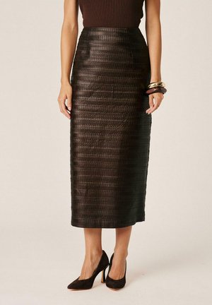 Mujer con falda lápiz midi marrón oscuro texturizada, top sin mangas oscuro, tacones altos negros y pulseras doradas y negras en la muñeca.
