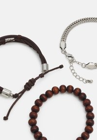 Trois bracelets : un en cuir marron avec des accents en argent, un en chaîne argentée, et un en perles en bois marron foncé, présentant des textures variées.