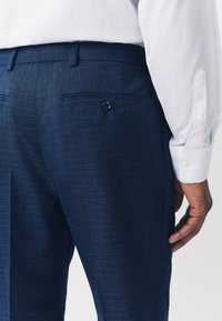 Blaue strukturierte Anzughose mit einer Seitentasche und Knopfdetails, kombiniert mit einem weißen Langarmshirt, das die Rückansicht und die Handplatzierung zeigt.