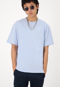 Camiseta de algodón azul claro con un corte relajado, mangas cortas y cuello redondo. Llevada con un collar de doble cadena y pantalones oscuros.