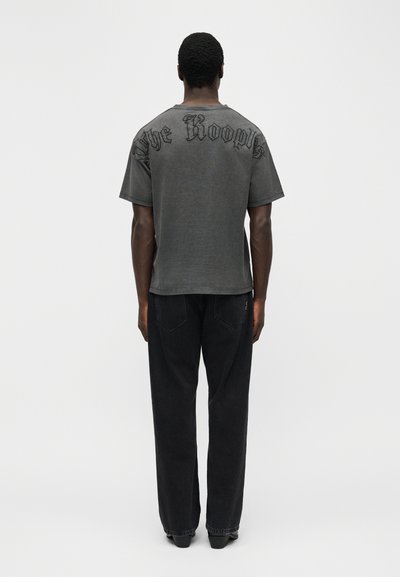 Άνδρας που στέκεται με την πλάτη προς τον θεατή, φοράει ένα ξεθωριασμένο μαύρο μπλουζάκι με το κείμενο "The Kooples" στους ώμους και χαλαρά μαύρα τζιν.