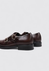 Zapatos de cuero marrón con acentos de doble hebilla y detalles de costura. Presentan una puntera redondeada y un tacón bajo negro. Textura suave.