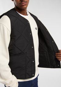 Chaleco negro acolchado con botones a presión, que cuenta con bolsillos laterales y una textura suave, superpuesto a un sudadera blanca.