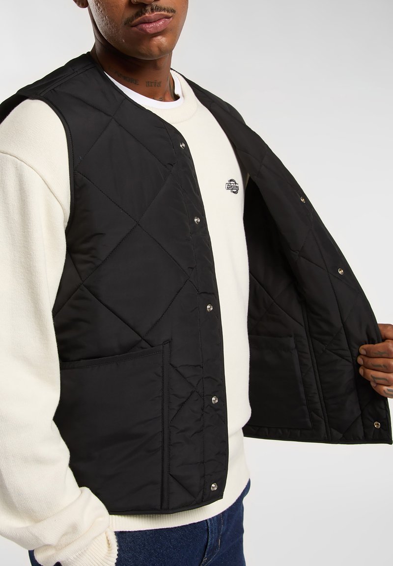 Sort quiltet vest med trykknapper, der har sidelommer og en glat tekstur, lagd over en hvid sweatshirt.