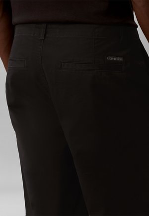 Prim-plan cu pantaloni negri cu două buzunare cusute la spate, având o etichetă mică Calvin Klein deasupra buzunarului drept.