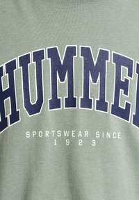 Grön sweatshirt med "HUMMER" i stora marinblå bokstäver och "SPORTSWEAR SINCE 1923" i mindre vit text nedanför. Mjuk bomullsmaterial.