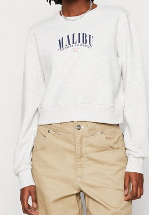 Personne portant un sweat-shirt gris clair avec le texte « MALIBU Southern California » et un petit motif de fleur, associé à un pantalon beige.