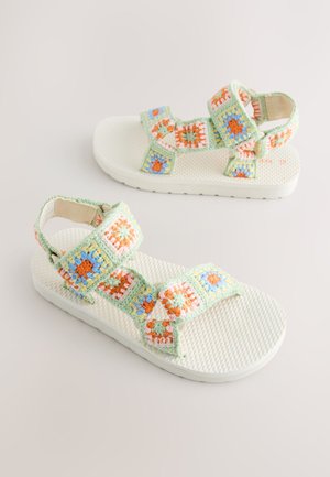 Zaļas un baltas sandales ar tamborētas stila siksnām, kurās ir oranži, zili un rozā ziedu raksti. Teksturēta balta zole.