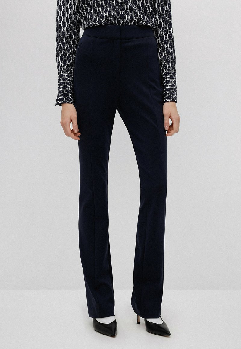 Pantalons tailleur navy avec une coupe droite, présentant une texture lisse et une taille haute. Associés à des chaussures noires à bout pointu.