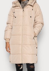 Beige pufferjakke med hætte, der har store forlommer, lynlås-lukning, elastiske manchetter og snoretræk i kanten. Glat, quiltet tekstur.