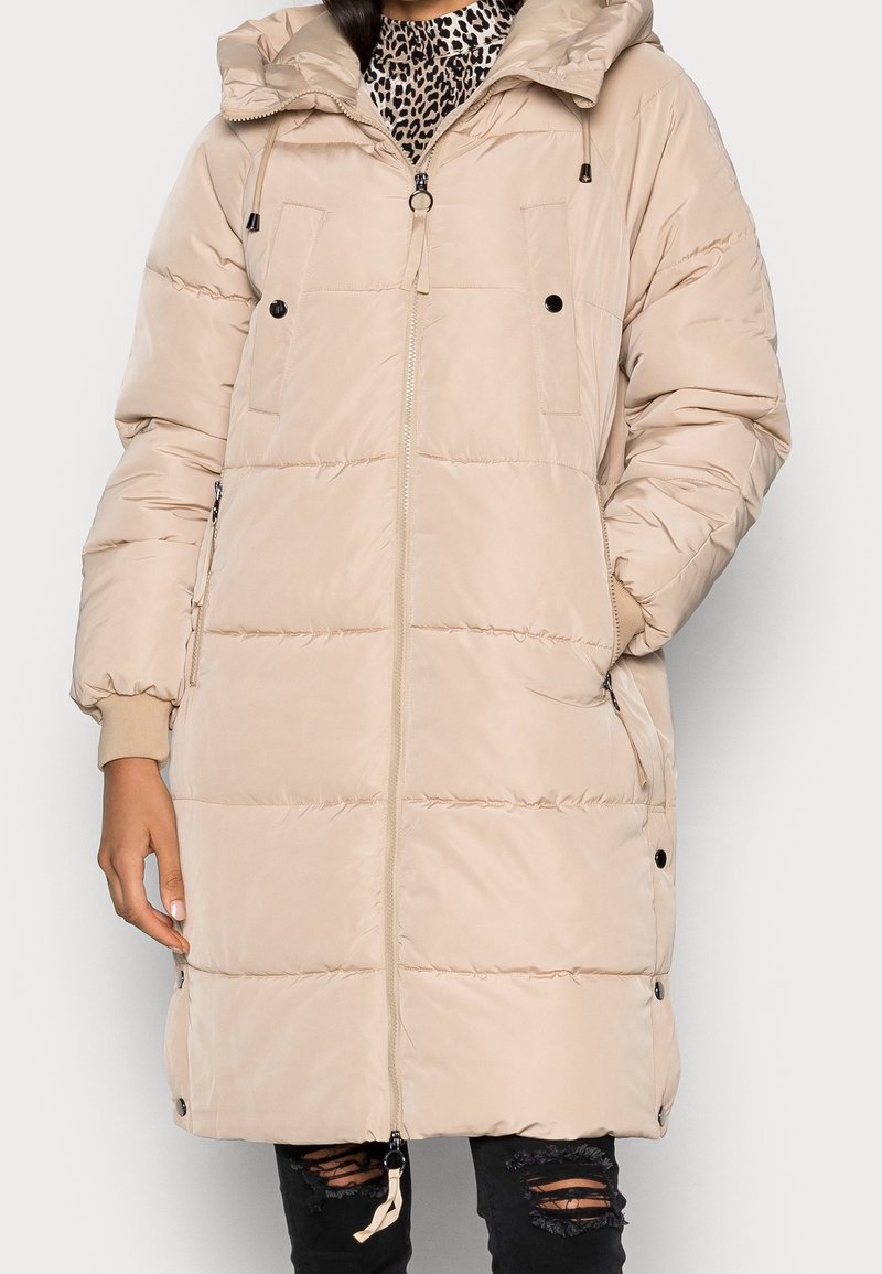 Beige pufferjakke med hætte, der har store forlommer, lynlås-lukning, elastiske manchetter og snoretræk i kanten. Glat, quiltet tekstur.