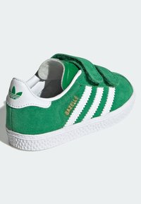 Green suede sneaker with white stripe accents and a textured rubber sole. Features a Velcro strap and embossed logo on the heel.