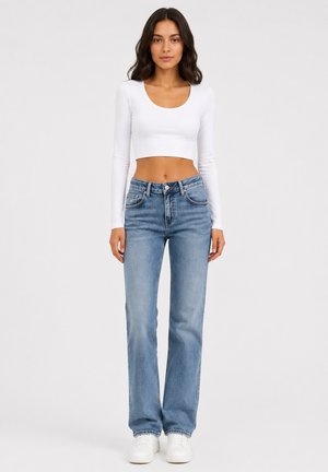 Kvinde iført en hvid langærmet cropped top, blå høj talje jeans og hvide sneakers, der står mod en ensfarvet baggrund.