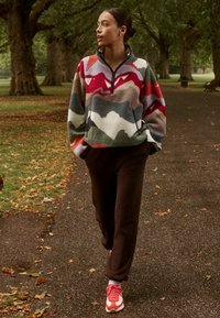 Pullover in felpa con un motivo a onde multicolore, dotato di collo con zip a metà; abbinato a pantaloni marroni e sneaker rosse e bianche.