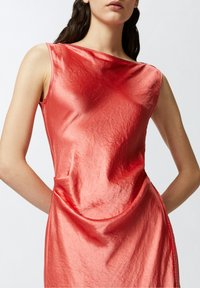 Koralska satenska haljina bez rukava s drapiranim dizajnom, koja se ističe zakrivljenim šavom i glatkom, lagano naboranom teksturom.