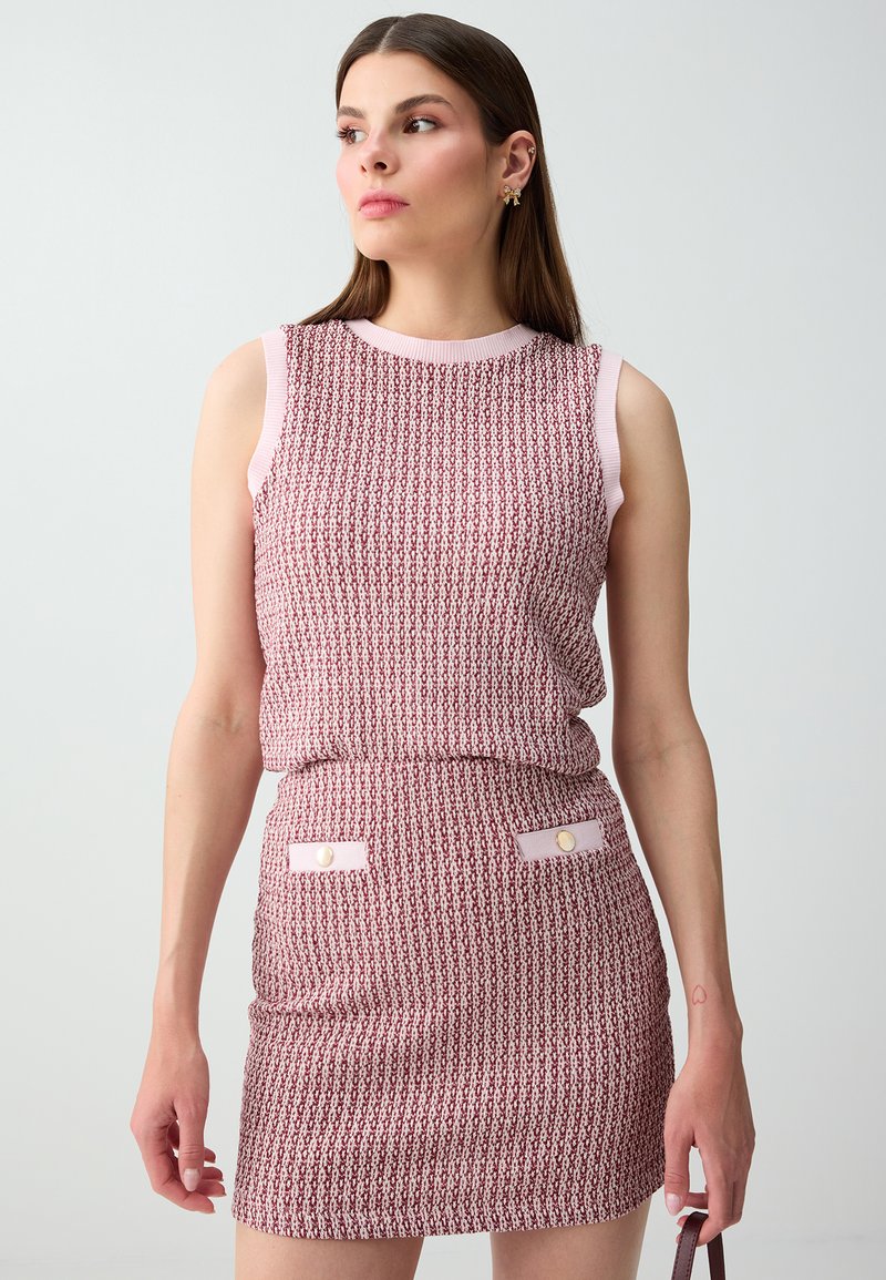 Femme portant un haut sans manches rose et rouge à motifs et une jupe assortie avec liseré rose et détails de boutons dorés, debout devant un fond uni.