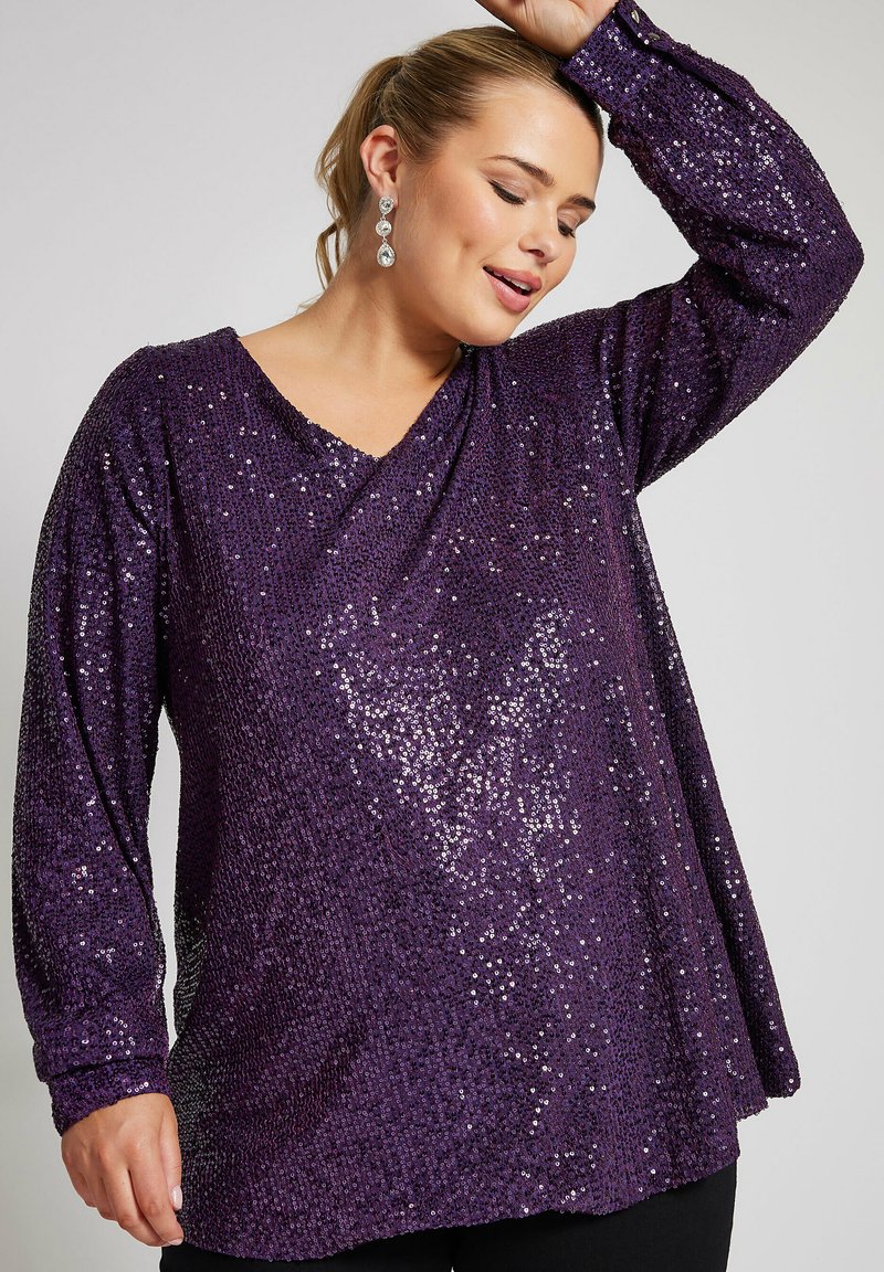 Lila paljetttopp med V-ringning, långa ärmar och en lös passform. Paljetterna skapar en strukturerad, reflekterande yta.