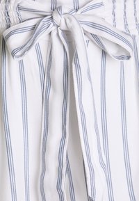 Witte stof met blauwe verticale strepen, voorzien van een geplooide tailleband en een gestrikt strikdetail aan de voorkant. Zachte textuur.