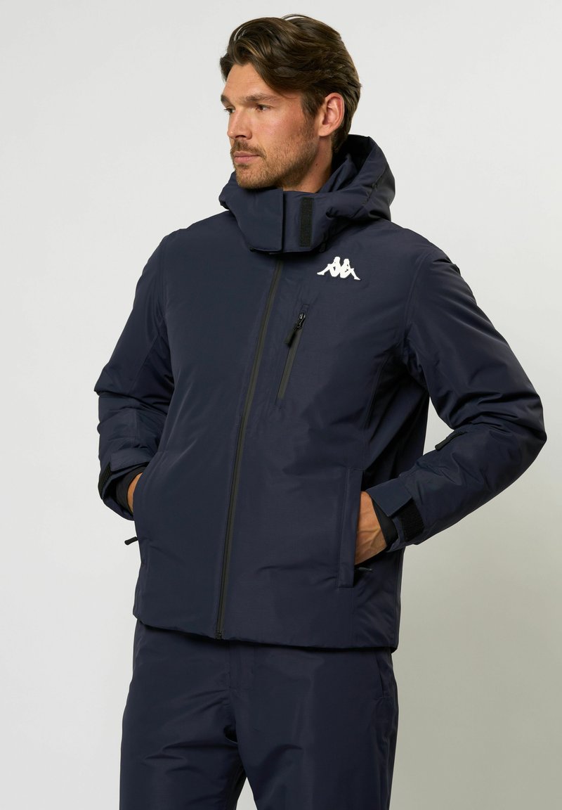 Marine isoleret jakke med høj krave, lynlåslukning foran og logodetalje. Inkluderer lommer og tætsiddende manchetter for varme og beskyttelse.