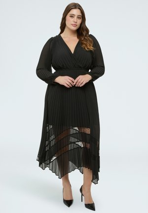 Frau, die ein schwarzes, langärmliges, plissiertes Midikleid mit transparenten Einsätzen und spitzen schwarzen High Heels trägt, steht vor einem schlichten Hintergrund.