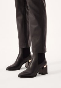 Botines negros con punta afilada, hechos de cuero suave. Lados elásticos acanalados y un tacón de bloque texturizado con un acento dorado.