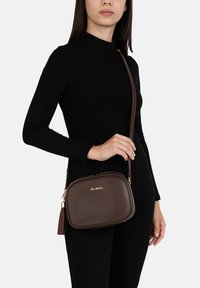Borsa a tracolla in pelle marrone con finitura testurizzata, chiusura con zip dorata e dettaglio a frange, tenuta da una persona che indossa un top nero.