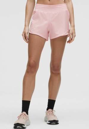 Woman wearing light pink athletic shorts, black crew socks, and pink running shoes standing against a neutral background.
