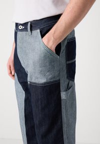 Denim pants featuring a patchwork design with light and dark blue tones, contrasting textures, and side pockets for functionality.