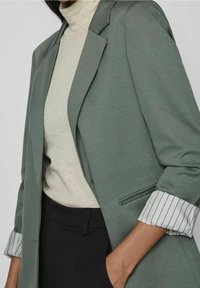 Zelené sako s texturovaným povrchem, s jedním knoflíkem a proužkovanými manžetami, nošené přes světlý rolák a černé kalhoty.