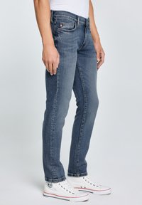 Mężczyzna ubrany w dopasowane niebieskie dżinsy i białe wysokie trampki, stojący na tle jasnego, jednolitego tła.