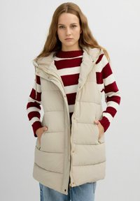 Chaleco puffy beige con capucha, que cuenta con cierre de cremallera y bolsillos laterales. El modelo lleva puesto un suéter a rayas rojas y blancas debajo.