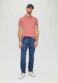 Rosa Poloshirt mit Kragen, kombiniert mit blauen Jeans und weißen Sneakers. Das Modell trägt eine marineblaue Kappe und steht vor einem schlichten Hintergrund.