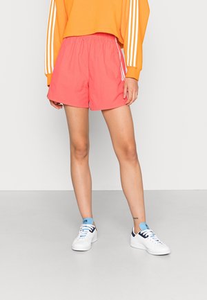 Persoon draagt koraalkleurige sportshorts, een oranje longsleeve met witte strepen en witte sneakers met lichtblauwe accenten.